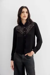 Camisa negra de mangas largas con cuello camisero y botones metálicos. Presenta tachas termofijadas en el frente y espalda.