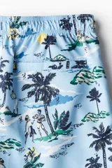 Short de baño celeste con estampado de palmeras, personas y paisajes playeros. Tiene elástico en la cintura y cordón de ajuste.