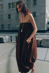 Vestido largo marrón de tejido satinado, con escote recto, tirantes finos extraíbles, bolsillos laterales ocultos y bajo asimétrico acabado en globo.