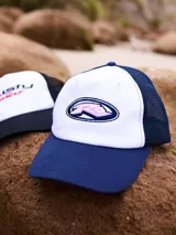 Gorra de lona blanca con visera azul marino y malla trasera azul marino. Presenta un logo bordado en el frente con letras rosadas y contorno azul marino.