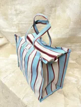Bolso tipo tote grande con rayas verticales en celeste, blanco y bordó. Tiene doble asa corta en celeste con borde bordó, cierre superior y bolsillo interno.