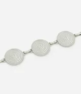 Cinturón de metal plateado, estilo cadena, compuesto por discos circulares con diseño de espiral concéntrica. Se ajusta mediante una cadena extensible con cierre de mosquetón.