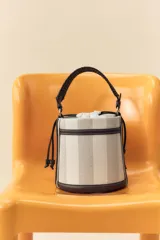 Bolso tipo bucket cilíndrico a rayas verticales en tonos beige y blanco, con asa de mano trenzada color marrón y asa de hombro extraíble. Incluye bolso de tela interior y pespuntes a contraste.