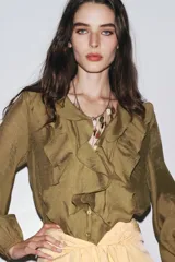 Camisa color verde oliva de tela traslúcida, con escote en V y volantes en el frente y cuello. Mangas largas abullonadas con puños elásticos.