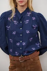 Camisa azul marino con estampado floral en color lila, cuello mao con volantes, cierre frontal de botones y mangas largas abullonadas con puños elásticos.