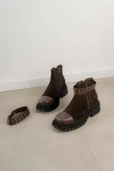 Bota corta de cuero gamuzado color marrón chocolate, con punta cuadrada y suela track. Presenta una correa con hebilla desmontable y cierre lateral con cremallera.