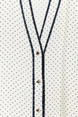 Camisa blanca con lunares negros, cuello redondo y escote pico. Sin mangas, con detalle de ribete negro en el escote y cierre frontal con botones.