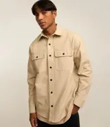 Camisa beige de manga larga confeccionada en sarga, con cuello inglés, abotonada al frente y en los puños. Presenta dos bolsillos con solapa en el pecho.