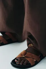 Sandalia plana de serraje color marrón, con dos tiras cruzadas en la parte delantera y una tira ancha con hebilla metálica ajustable.