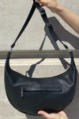 Cartera tipo bandolera de cuero sintético color celeste, con forma de media luna y tachas metálicas redondas plateadas en el frente. Posee correa ajustable y placa metálica con logo en la parte superior.