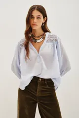 Camisa de algodón color celeste, con escote en V, cierre parcial de botones y detalles de encaje en los hombros. Mangas largas abullonadas con puños elásticos.