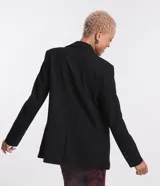 Blazer negro de sastrería con corte alargado, cuello con solapa, manga larga, botón delantero, bolsillos fake y cadena decorativa en la solapa.