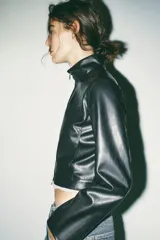 Cazadora corta negra de efecto piel, con cuello subido y manga larga. Tiene bolsillos delanteros con cremallera metálica y cierre frontal con cremallera.