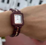 Reloj de pulsera con caja cuadrada y correa de eslabones de madera color borgoña. El dial es plateado con agujas y detalles dorados.