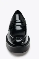 Zapato tipo mocasín de charol negro, con detalle de antifaz en el empeine y punta redonda.