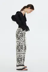Pantalón largo de viscosa color blanco con estampado de flores y figuras geométricas en negro. Tiene tiro alto, cintura elástica con cordón ajustable, bolsillos laterales y corte holgado.