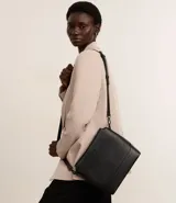 Bolso mediano tipo postman color negro con textura en relieve de líneas horizontales. Tiene correa larga regulable, cierre con cremallera y forro de poliéster.