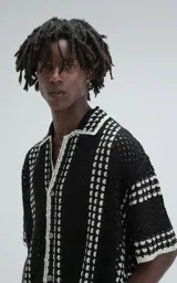 Camisa de manga corta color negro, tejida en punto abierto (símil crochet) con detalles en blanco. Tiene cuello tipo solapa, botones frontales y un calce oversize.