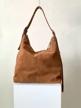 Bolso tipo hobo color tostado, hecho de cuero con flecos largos en el frente y dos bolsillos con cierre. Tiene una manija regulable con hebillas metálicas.
