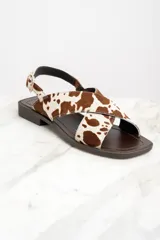 Sandalias bajas con tiras cruzadas de cuero con estampado de vaca en tonos marrón y blanco. Tira trasera con hebilla dorada y suela plana color marrón.