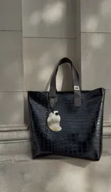 Bolso tote negro con textura tipo piel de cocodrilo, doble asa corta y dije colgante.