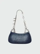 Bolso de hombro Marc Jacobs modelo The Shoulder Bag Charm, confeccionado en denim cepillado azul. Presenta una correa de cadena plateada con dijes colgantes, incluyendo una estrella y un alfiler de gancho. El bolso tiene forma curva y un cierre superior.