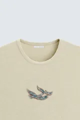 Camiseta sin mangas color arena con cuello redondo y estampado de ave en el pecho.
