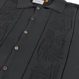 Camisa de punto de manga corta color negro, con cuello tipo camp y botones frontales. Presenta detalles de bordados tonales con motivos botánicos y franjas verticales texturizadas.