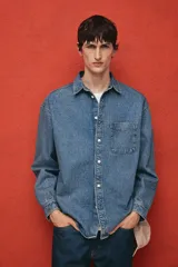 Camisa de denim azul de algodón rígido, corte holgado, cuello inglés, tapeta clásica con botones blancos, bolsillo de parche en el pecho, canesú en la espalda, mangas largas con puños abotonados y bajo levemente redondeado.