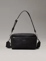 Cartera tipo cámara convertible Calvin Klein negra, con textura monograma en el bolsillo frontal y logo metálico plateado. Incluye dos correas desmontables: una ancha ajustable para cruzar y una fina para llevar al hombro.
