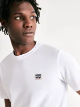 Camiseta blanca de algodón con cuello redondo y mangas cortas. Estampa rectangular con borde negro que contiene la frase "NATURAL ATMOSPHERE" en letras mayúsculas y una imagen de montañas y mar.