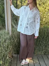 Camisa blanca de broderie con diseño floral, corte oversize, cuello clásico, cierre frontal con botones y mangas largas con puños abotonados.