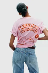 Camiseta rosa de algodón con efecto lavado, corte regular, cuello redondo y mangas cortas. Estampado frontal con la frase "Puerto Rico Vuela Conmigo".