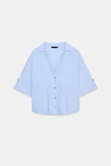 Camisa de lino color celeste claro, con cuello solapa, escote pico, manga corta con vuelta y dos bolsillos de plastrón en el delantero. Cierre frontal con botones.