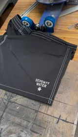 Short de baño negro de largo medio (18 pulgadas) con costuras reforzadas en hilo blanco contrastante. Presenta cordón de ajuste en la cintura y un pequeño logo estampado en la pierna derecha.