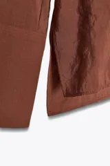 Camisa marrón de Zara Woman Collection confeccionada en hilatura con mezcla de lyocell. Tiene cuello redondo, manga larga acabada en puño, bajo asimétrico con aberturas laterales y cierre frontal con botones.
