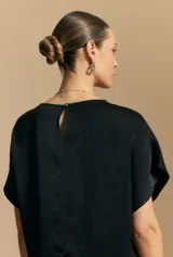 Top negro de manga corta con silueta amplia y caída fluida, confeccionado en un tejido liviano con leve brillo. Presenta un botón de nácar en la espalda con una pequeña abertura y mangas amplias tipo kimono.