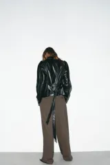 Chaqueta negra de efecto piel con cuello solapa, manga larga y trabillas en los hombros. Presenta bolsillos delanteros y cierre frontal con botones, ajustada a la cintura con un cinturón del mismo tejido.