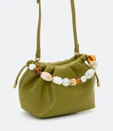 Bolso pequeño color verde oliva, desestructurado, con pinzas frontales y cierre de botón a presión. Incluye una correa de hombro extraíble y un asa de mano decorada con cuentas acrílicas variadas en tonos blancos, marrones y transparentes.