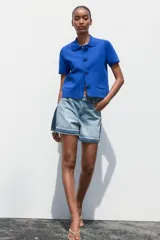 Chaqueta de punto azul con cuello solapa y manga corta. Tiene bolsillos delanteros de parche y cierre frontal con botones grandes con abalorios.