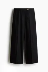 Pantalón sastrero tobillero negro de tiro alto con cierre y pretina prolongada con botón. Presenta bolsillos al bies, bolsillos traseros decorativos y piernas amplias con pliegues adelante y atrás.