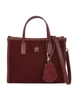 Cartera tipo tote bag de color borgoña, con acabado liso y detalles en cuero sintético del mismo tono. Presenta dos asas superiores y una correa ajustable extraíble. Incluye un colgante decorativo con el escudo de Hilfiger bordado y un dije metálico con el monograma TH.