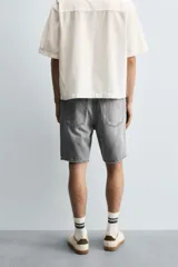 Bermuda de denim de algodón, color gris lavado, con corte relaxed fit y roturas visibles en la parte delantera de ambas piernas. Presenta cinco bolsillos y cierre frontal con cremallera y botón.