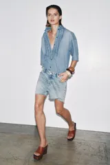 Camisa de denim celeste con cuello solapa y manga corta. Presenta detalle de volantes en el frente y bajo asimétrico con aberturas laterales.