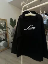 Buzo hoodie negro de corte oversize, con capucha, bolsillo frontal y estampa blanca en la espalda con la leyenda "Luxe" y "The Luxe Vip Club".