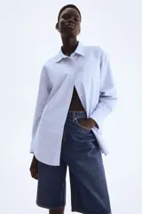 Camisa celeste y blanca a rayas verticales, de corte holgado, con cuello clásico, botones frontales, mangas largas con puños abotonados y bajo redondeado.