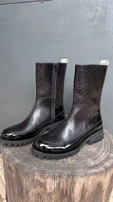 Bota de caña 3/4 color negro, con textura de cocodrilo en la parte superior y acabado charol en la puntera y el talón. Tiene cierre lateral y suela track.