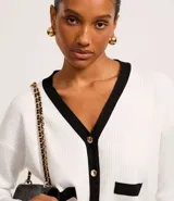 Cardigan negro de punto con escote en V, botones metalizados dorados, bolsillos delanteros falsos, cuello y borde contrastantes en blanco.