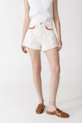 Short de denim color blanco roto con bordes deshilachados y detalles de crochet en color rojo en los bordes de los bolsillos delanteros.