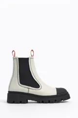 Botines Chelsea de cuero sintético blanco con puntera y suela track de goma negra. Elásticos laterales negros y tirador trasero naranja.